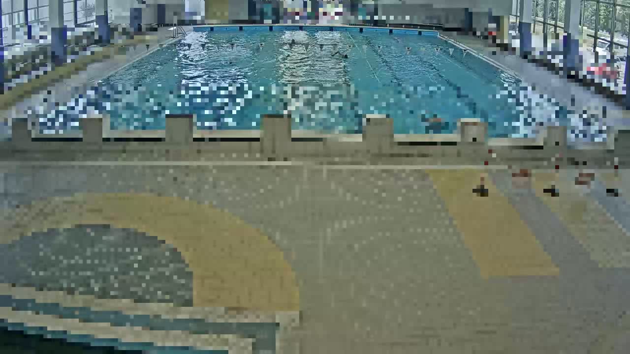 An indoor swimming pool with many people swimming in it, surrounded by a tiled floor and large windows.