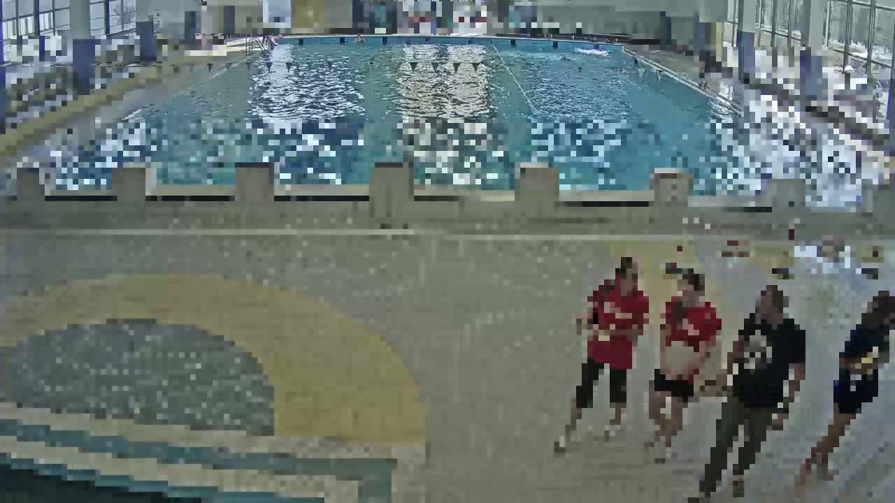 An indoor swimming pool with several people standing on the pool deck, near the side of a large, mostly empty pool.