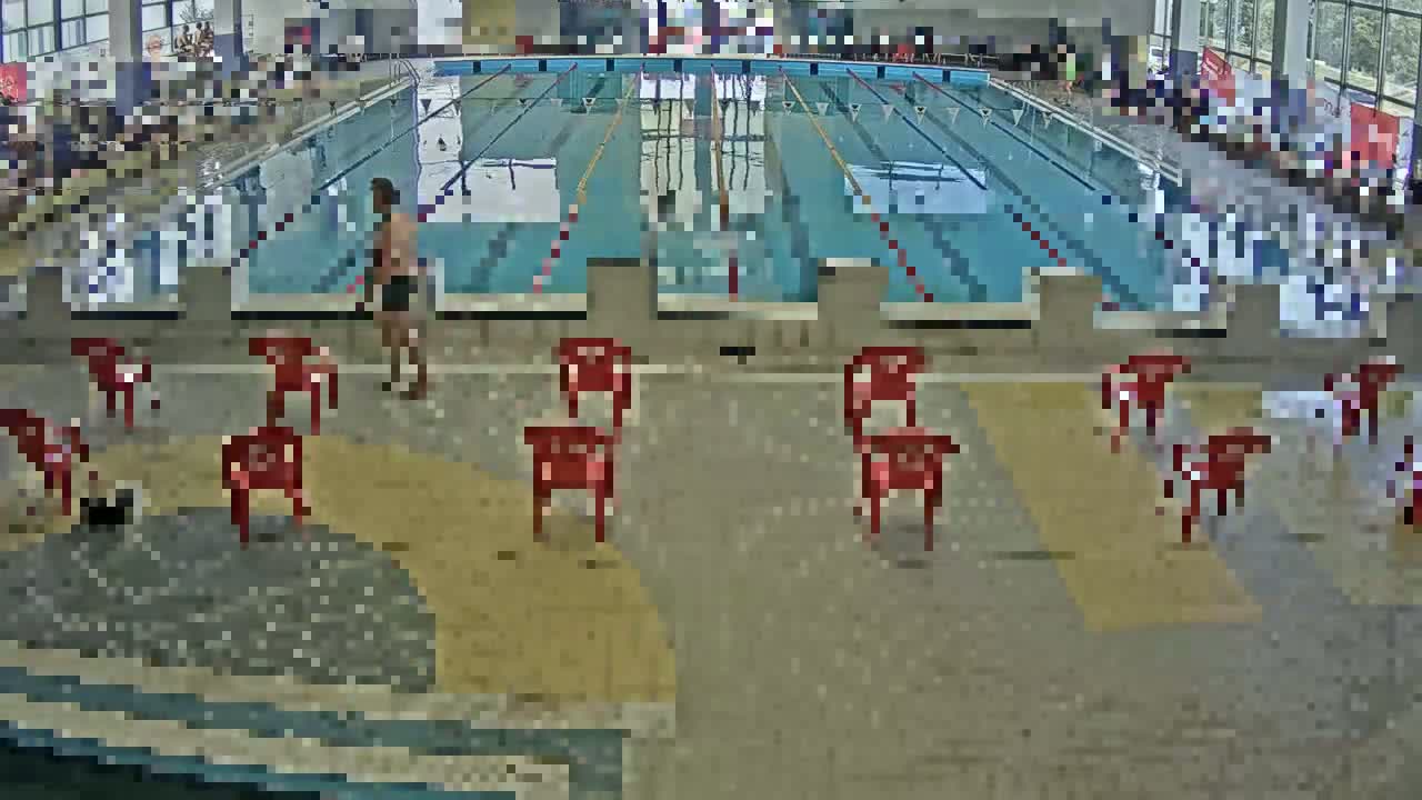 An indoor swimming pool with multiple lanes, a man standing near the side, and several red plastic chairs scattered around the pool deck.