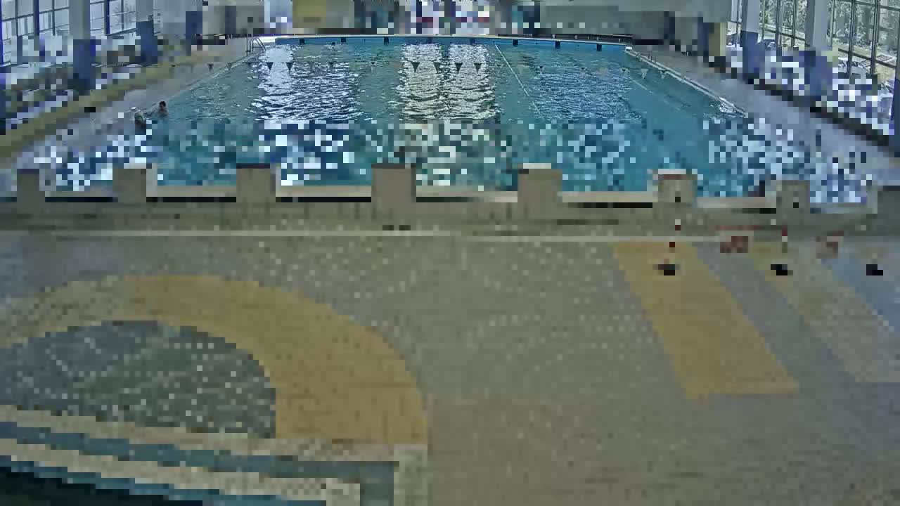 An indoor swimming pool with a few people in it is viewed from above, the pool area surrounded by a tiled floor.
