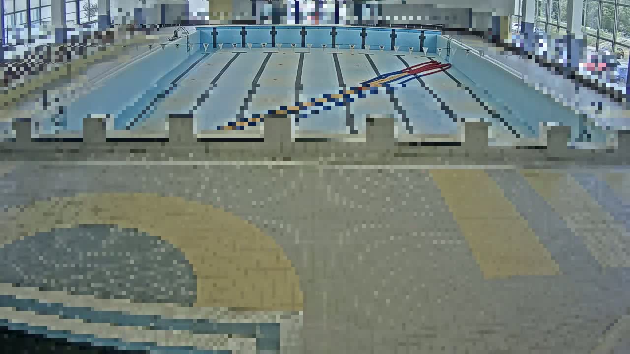 An indoor swimming pool with lane dividers and starting blocks is viewed from above, showing the pool deck and surrounding area.