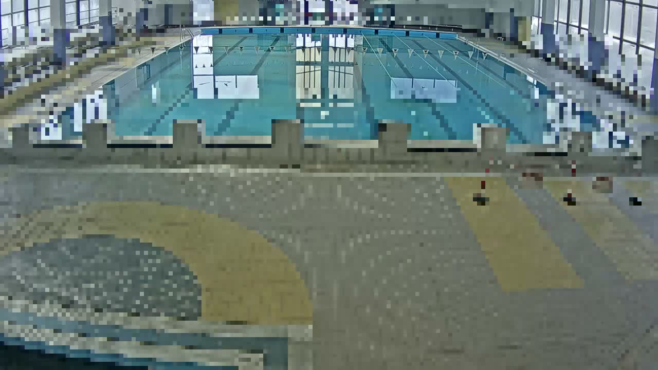 An indoor swimming pool with multiple empty lanes, marked by ropes and blue lines, is visible from an elevated perspective, surrounded by a tiled deck featuring yellow and gray patterns and illuminated by natural light from large windows.