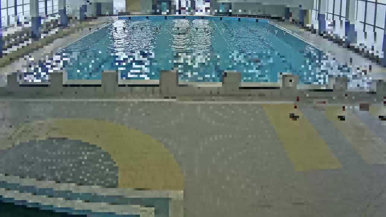 A brightly lit indoor swimming pool with multiple marked lanes, starting blocks, and patterned grey and yellow flooring in the foreground, illuminated by large windows along the back.