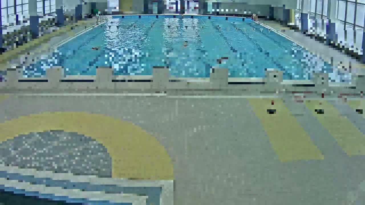 A brightly lit indoor swimming pool with multiple marked lanes, starting blocks, and patterned grey and yellow flooring in the foreground, illuminated by large windows along the back.