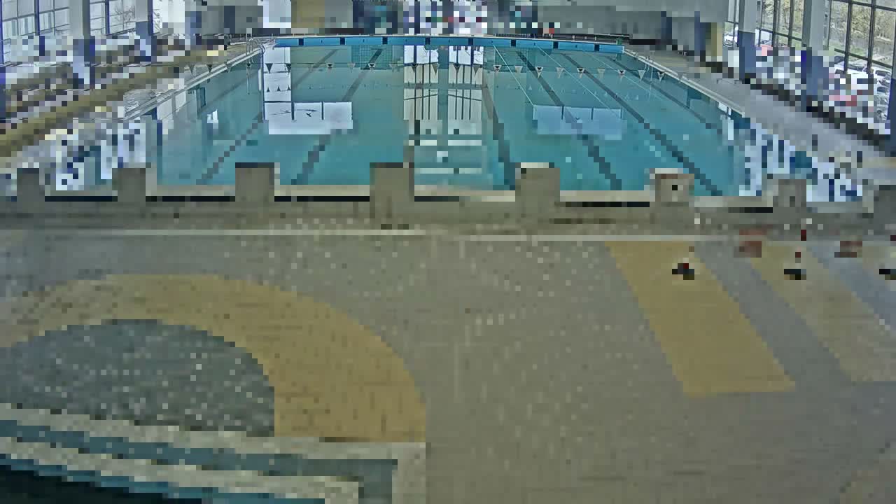 A brightly lit indoor swimming pool with multiple marked lanes, starting blocks, and patterned grey and yellow flooring in the foreground, illuminated by large windows along the back.