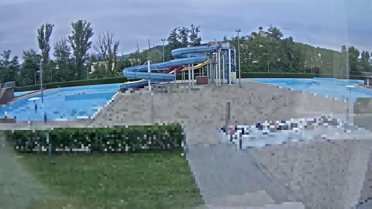 An outdoor swimming pool complex with water slides is visible under an overcast sky, with a hill and trees in the background.