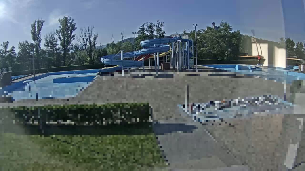 An outdoor swimming pool with water slides is surrounded by trees and shrubs under a mostly sunny sky.