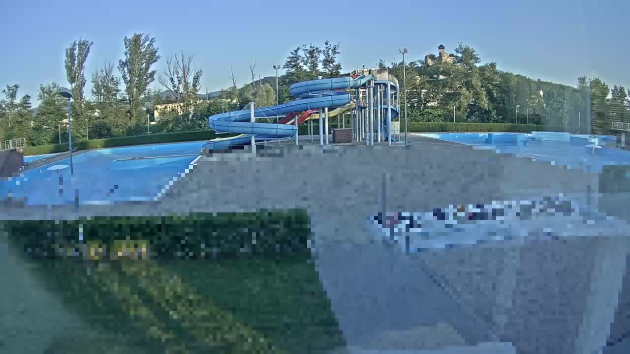 A swimming pool complex with two twisting water slides is visible under a clear, sunny sky, with a hill and trees in the background.