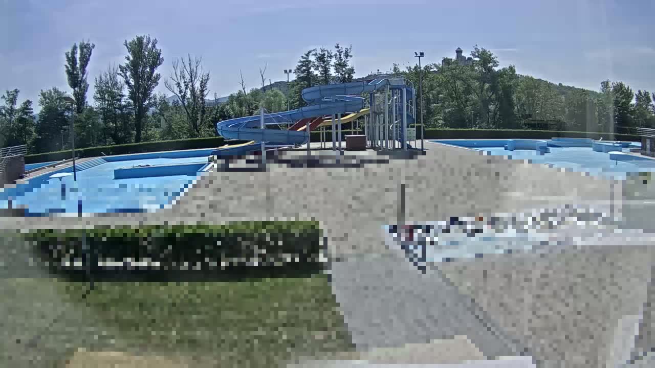 An outdoor swimming pool complex with water slides is situated on a sunny day near a tree-covered hill.