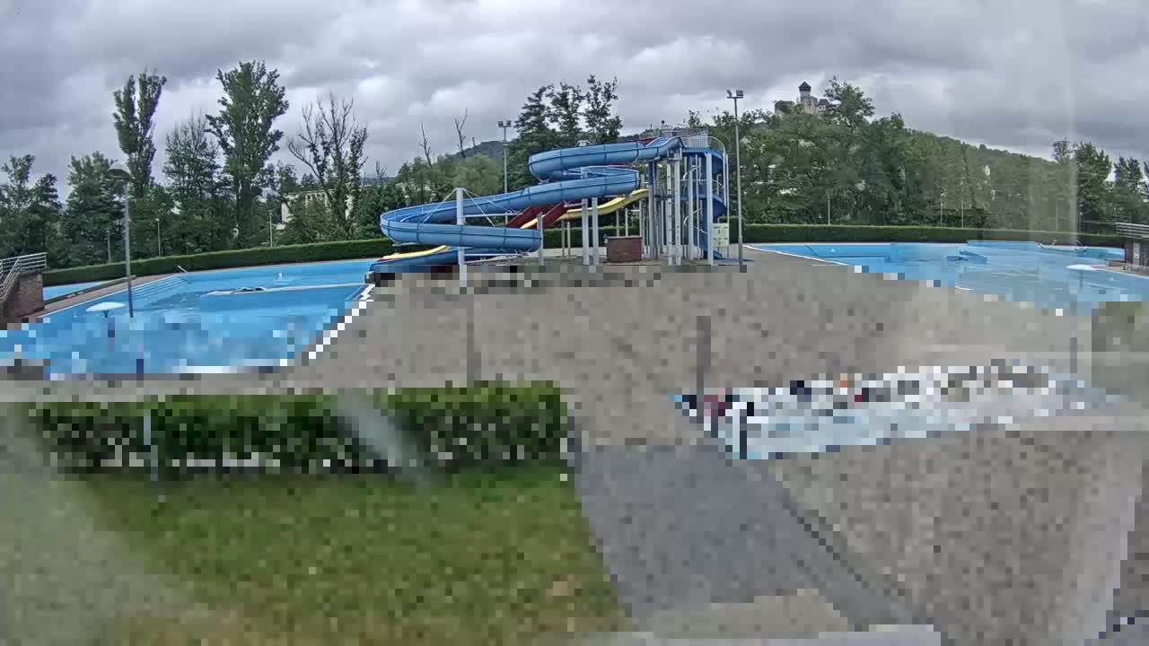 An outdoor swimming pool complex with two water slides sits under a cloudy sky, surrounded by greenery and a hill in the background.
