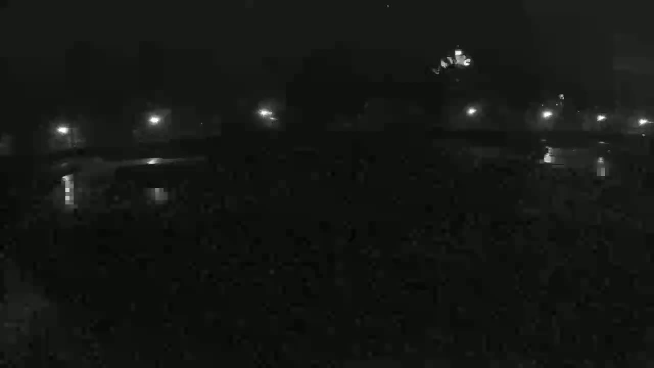 A nighttime, dimly lit outdoor scene shows several dark vehicles parked in a lot, illuminated by scattered streetlights under a dark, possibly overcast sky.