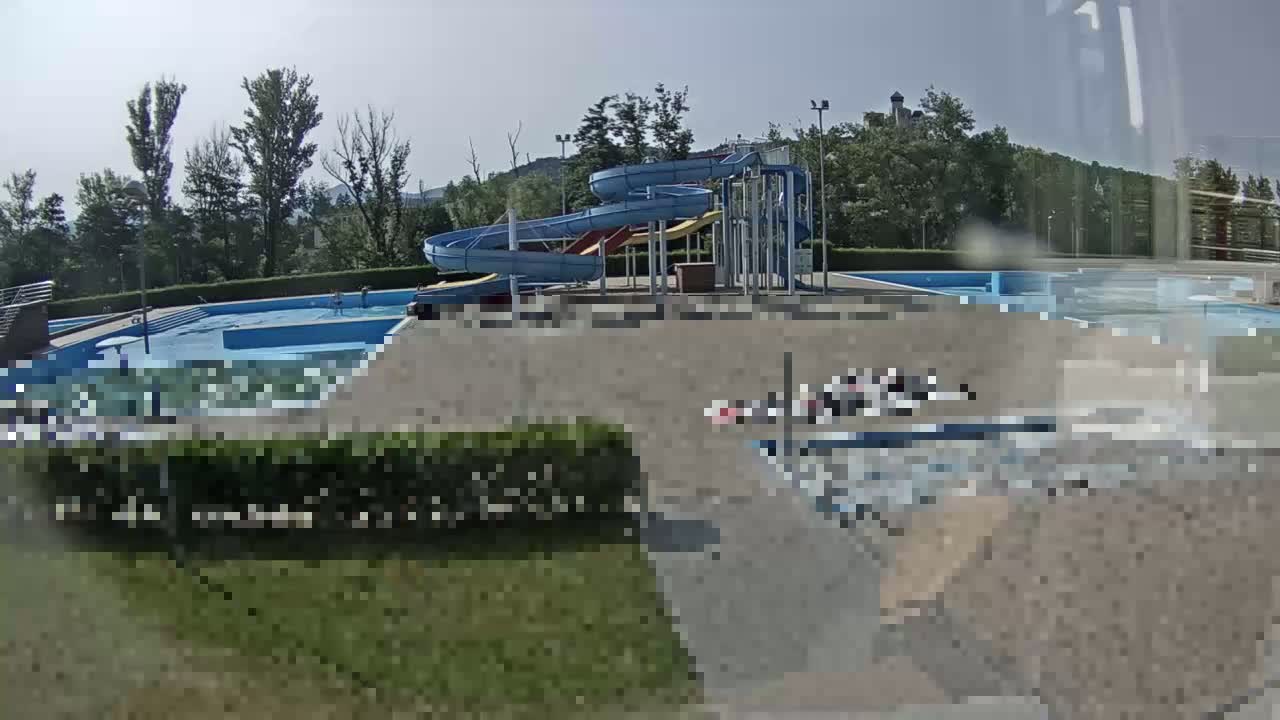 A sunny outdoor scene shows a swimming pool complex with multiple pools and a tall waterslide structure.