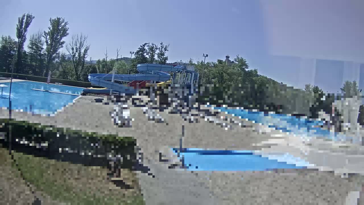 A sunny outdoor water park features a large swimming pool and water slides, surrounded by lounge chairs and trees.