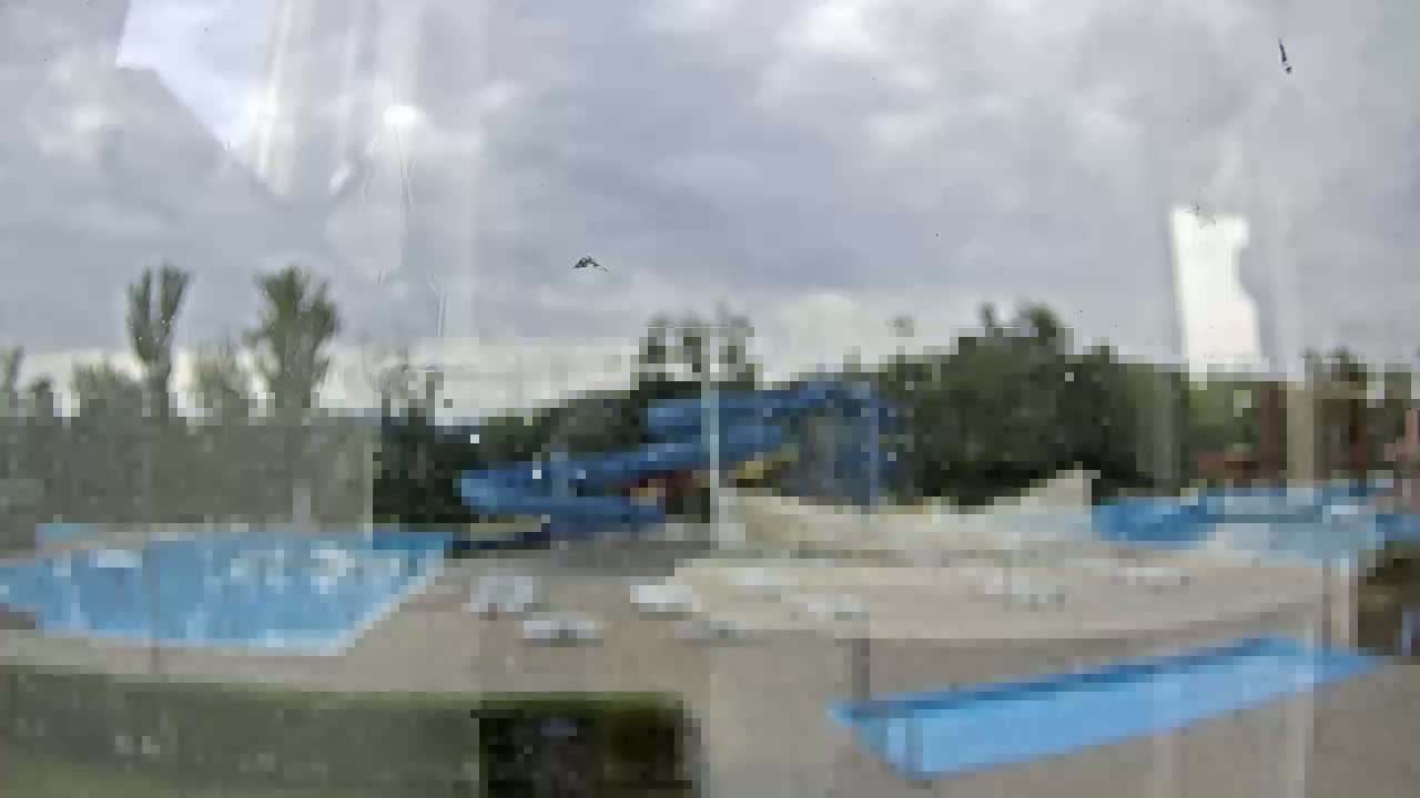 A view through a dirty window shows an outdoor water park with several pools and water slides under an overcast sky.