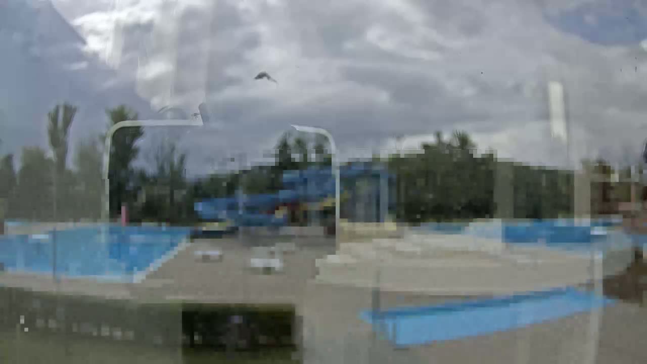 A view through a window shows an outdoor water park with multiple swimming pools under a cloudy sky.