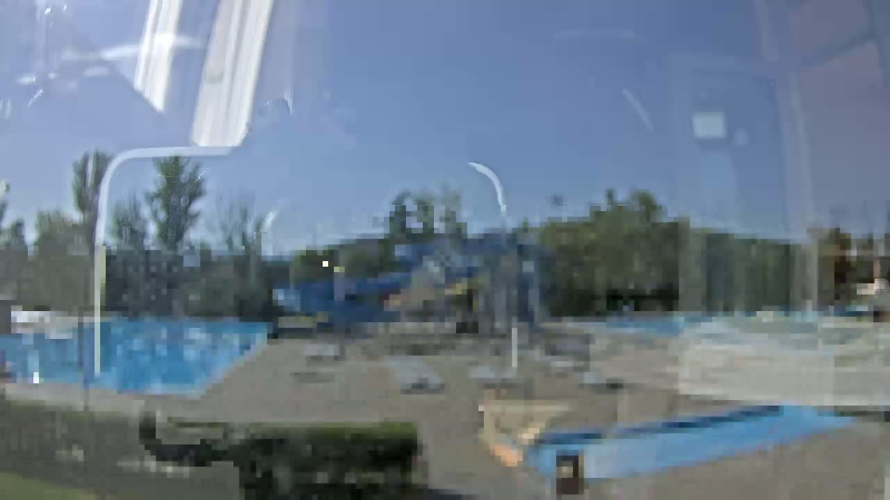 A blurry view through a window shows a swimming pool and surrounding area on a sunny day.