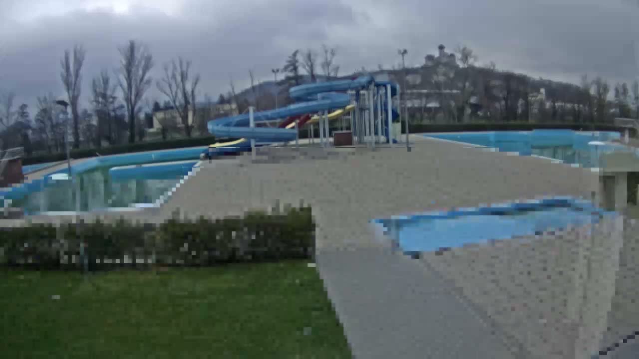 An outdoor water park featuring a large blue and yellow water slide complex, multiple swimming pools (some appearing empty or partially covered), concrete pathways, and bare trees in the background under a cloudy, overcast sky.