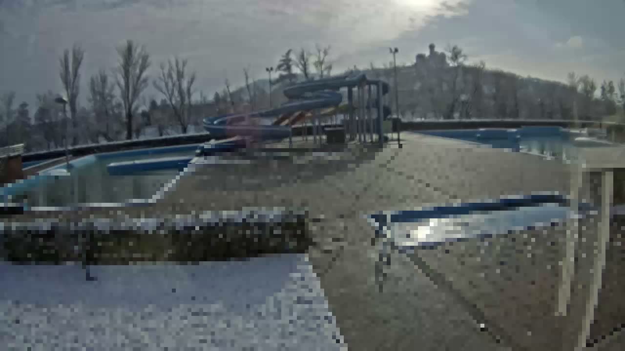An outdoor water park with a large blue water slide and multiple pools is seen covered in snow, surrounded by bare trees under a cloudy sky.