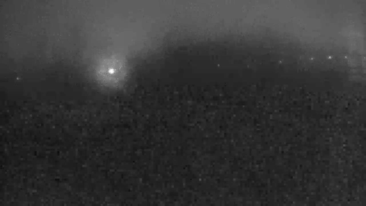 A heavily fogged outdoor scene at night reveals a prominent bright light on the left and a distant line of smaller lights stretching across the horizon, over a dark and indistinct foreground.