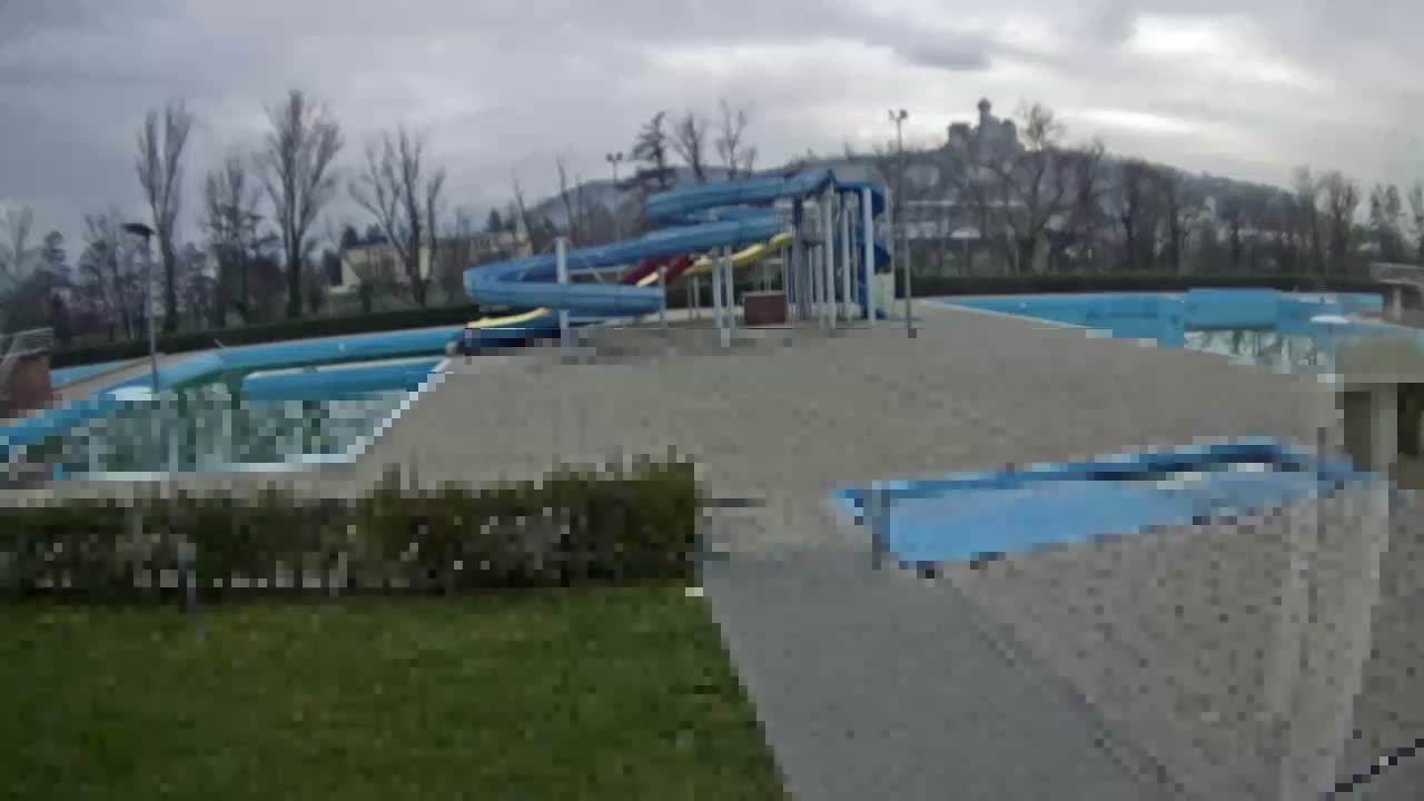 An empty outdoor swimming pool complex with multiple slides and bare trees stands under an overcast sky, with a distant building visible on a hill.