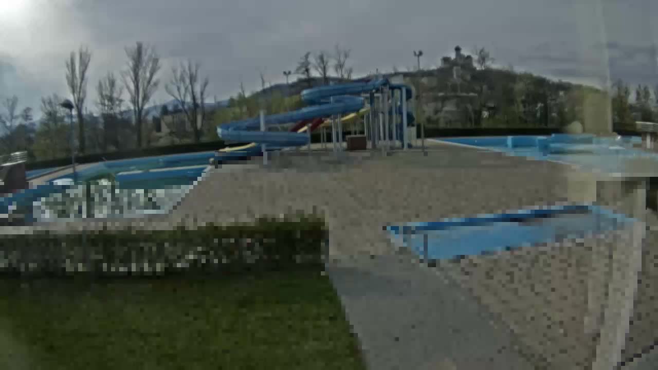 An empty outdoor swimming pool complex with multiple slides and bare trees stands under an overcast sky, with a distant building visible on a hill.