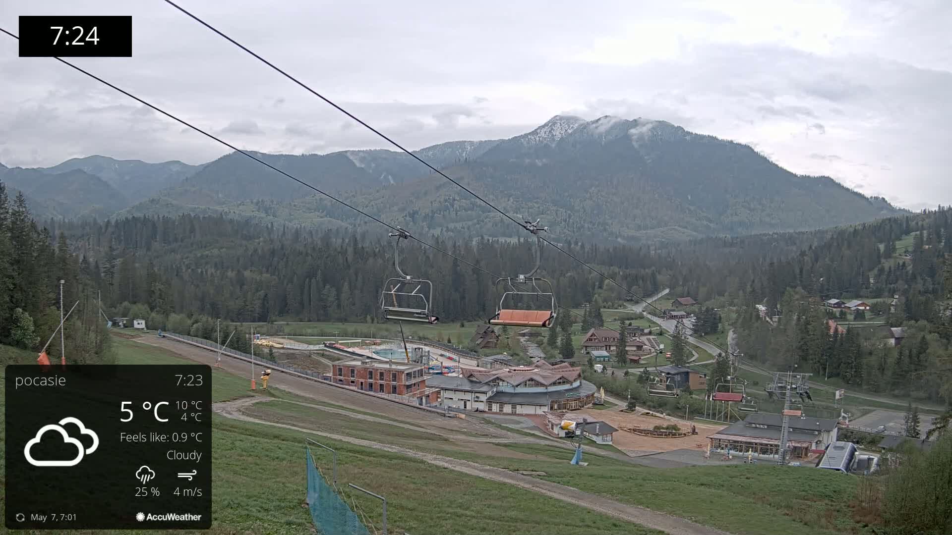 A cloudy, cool day at a ski resort shows an empty chairlift overlooking a village nestled in a valley below snow-capped mountains.