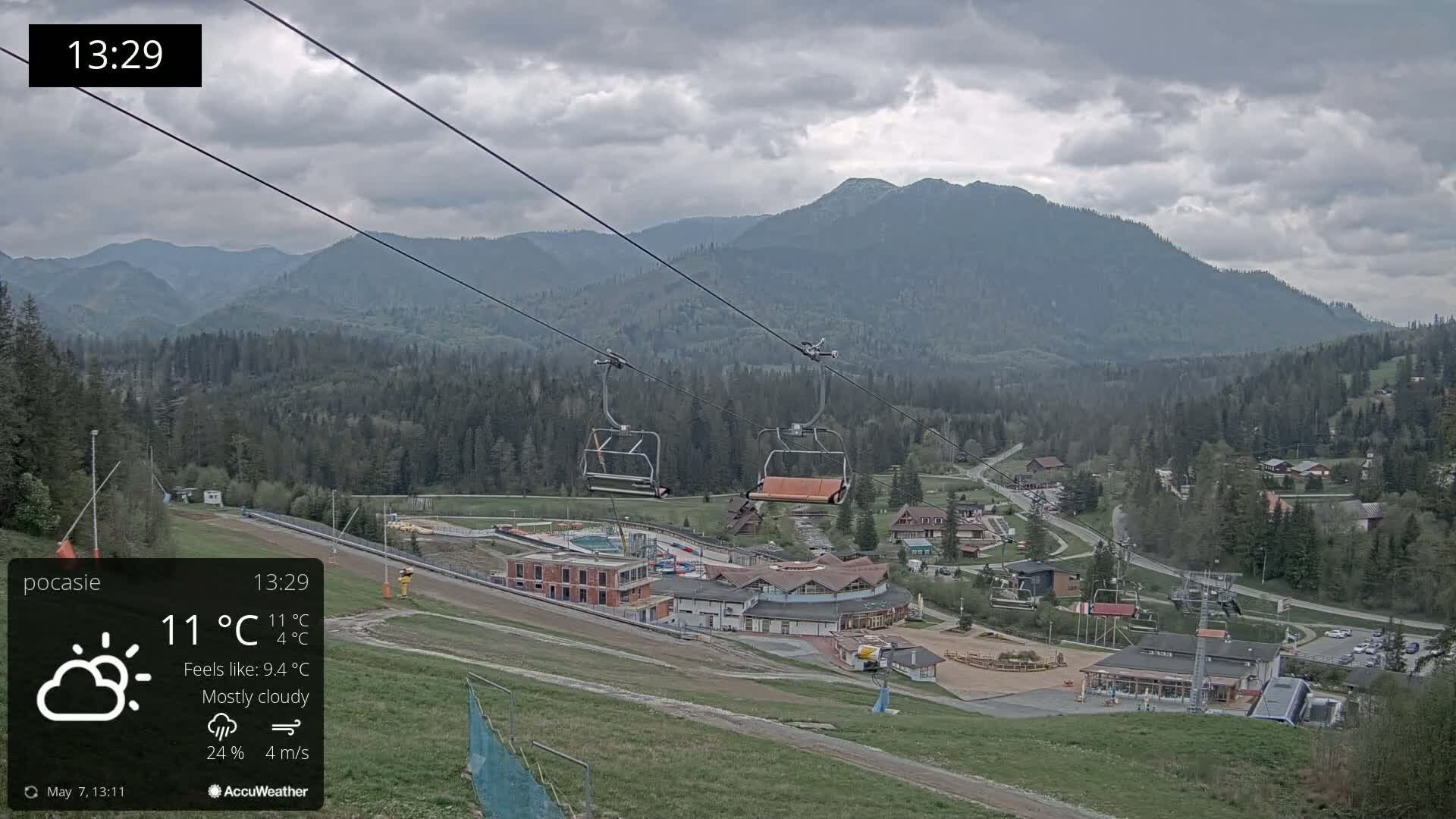 A mostly cloudy mountain vista shows an empty chairlift ascending towards a ski resort nestled among green hills and forests under a gray sky.