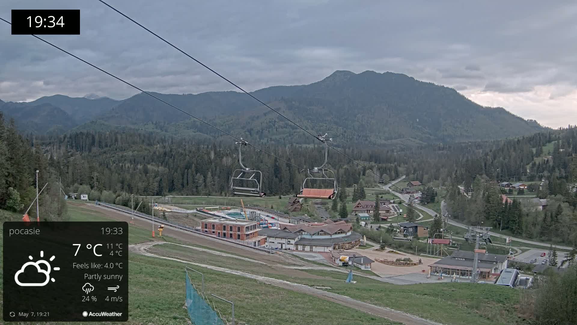 An empty chairlift ascends a mountainside overlooking a partly sunny resort town nestled among forested hills.