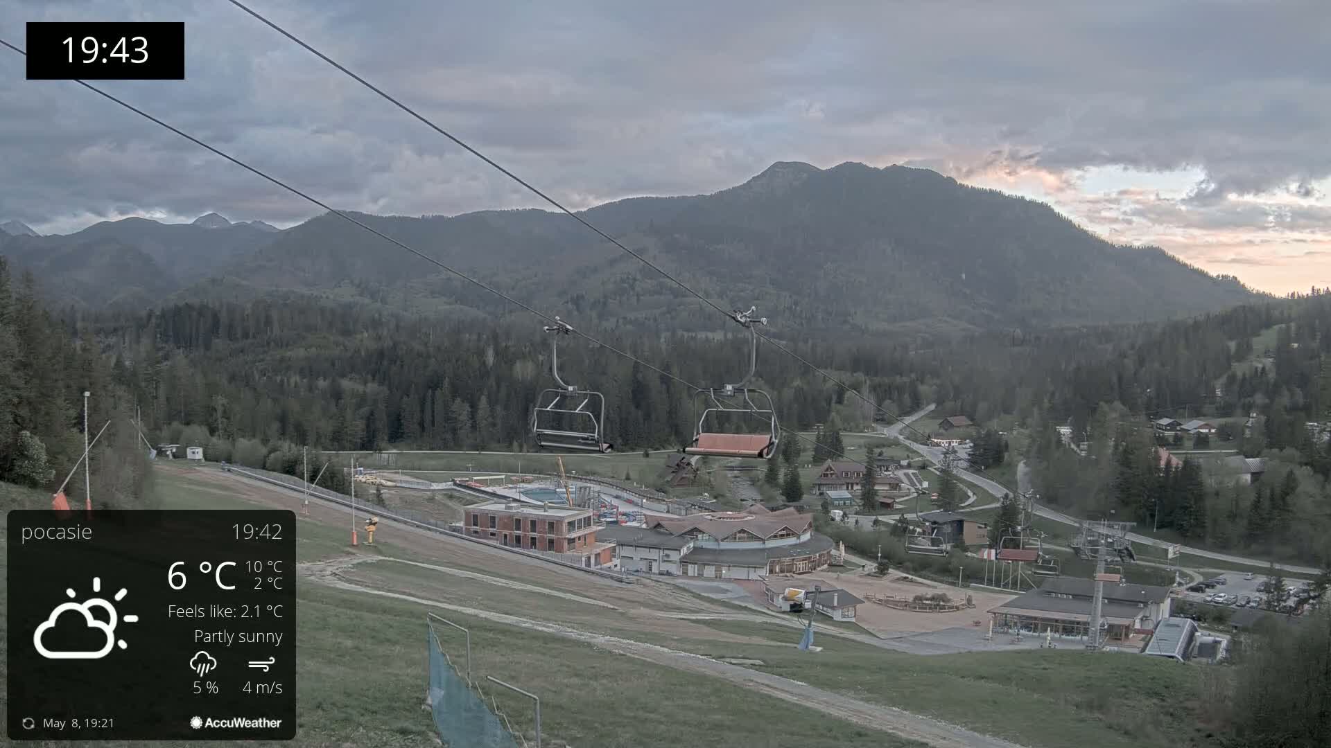 An empty chairlift ascends a mountainside overlooking a partly sunny village nestled among the foothills, under a partly cloudy sky.