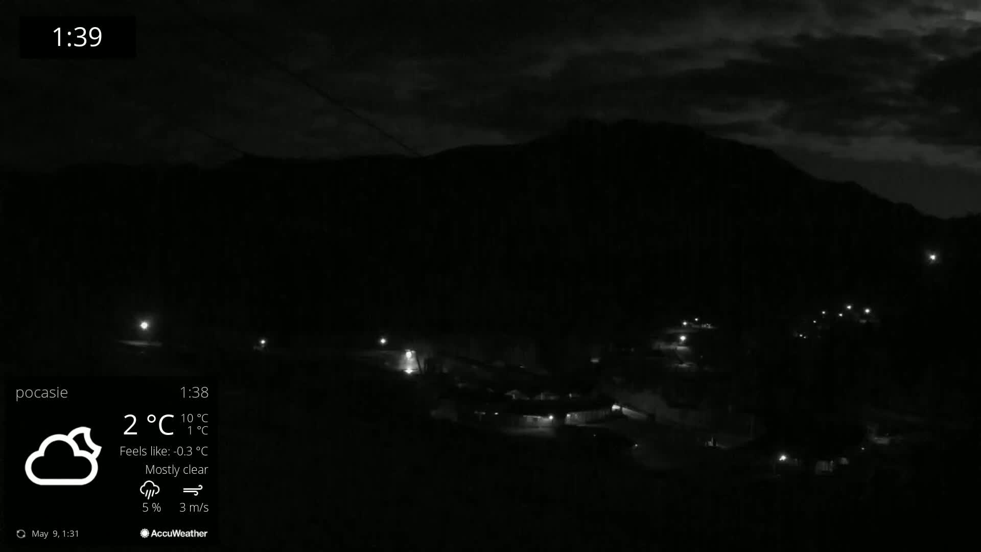 A nighttime, mostly clear view of a small town nestled below a dark mountain, illuminated by scattered lights.