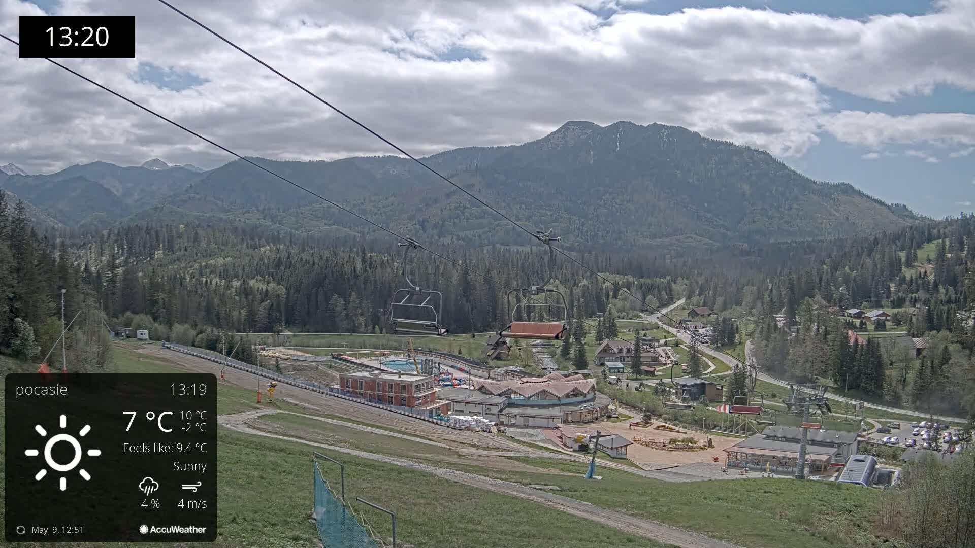 A sunny mountain resort with a chairlift and buildings is visible, nestled among green forests and mountains under a partly cloudy sky.