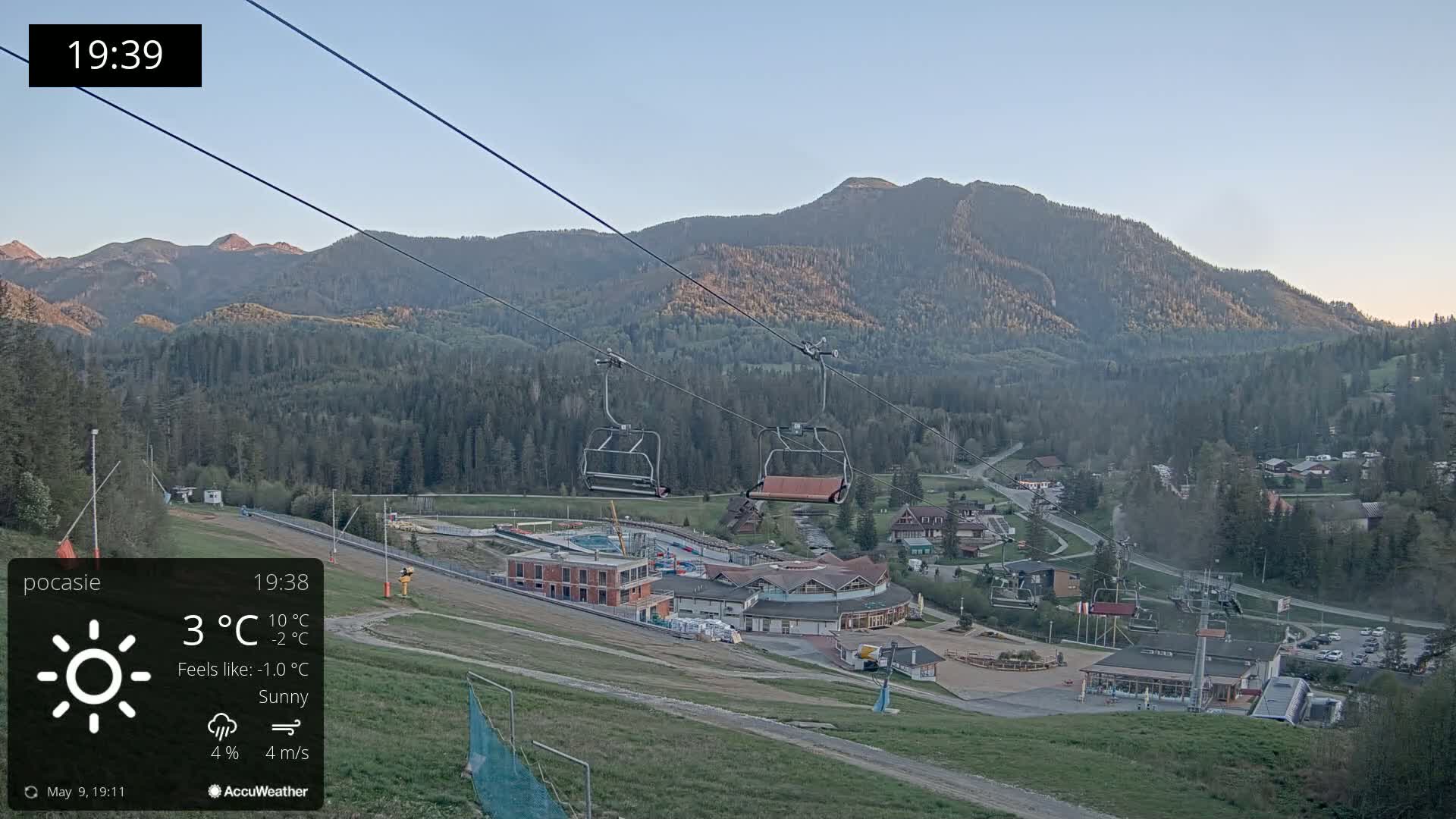 Under sunny skies with a light breeze, an empty chairlift ascends a mountainside toward a village nestled in a valley.