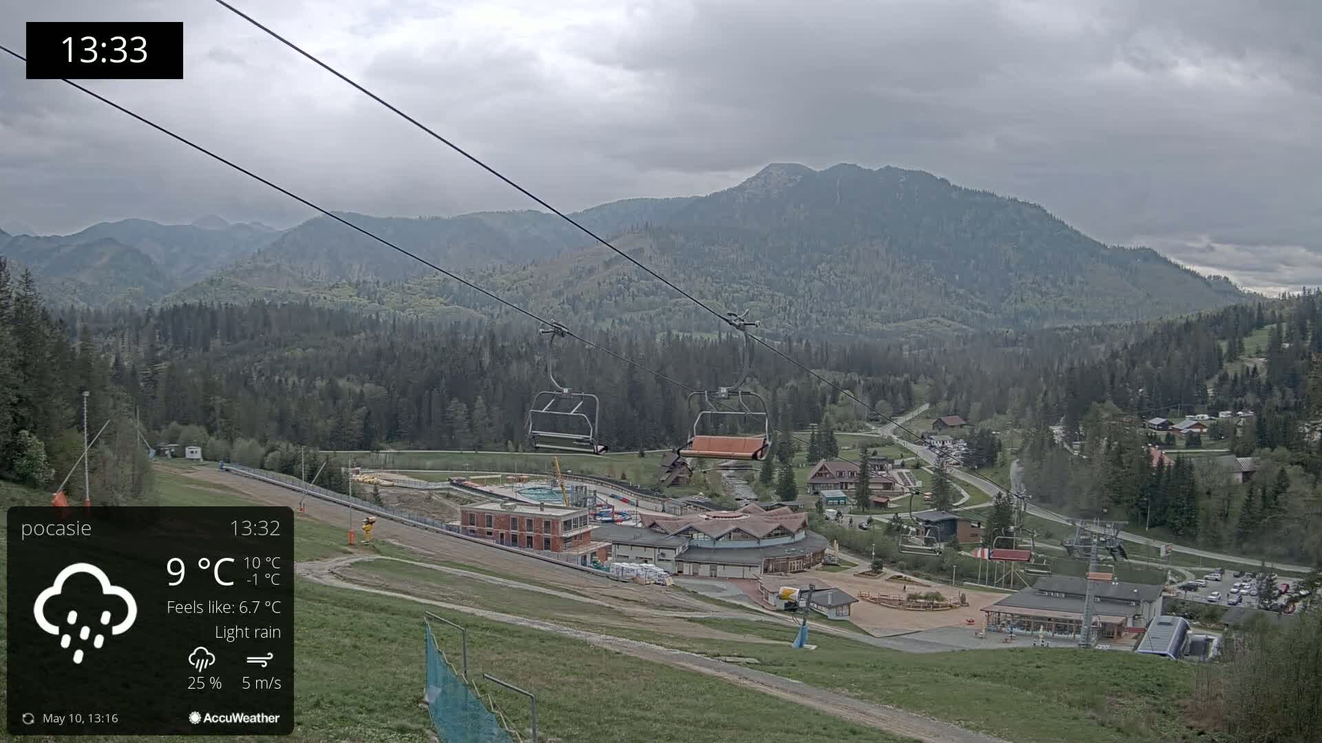 A chairlift ascends a mountainside overlooking a village under a light rain and overcast skies.