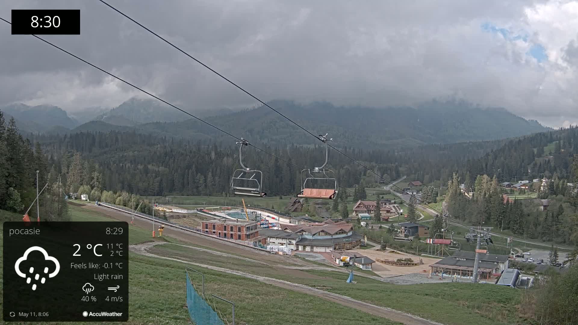 A chairlift ascends a mountainside overlooking a village and a water park under a cloudy sky with light rain.