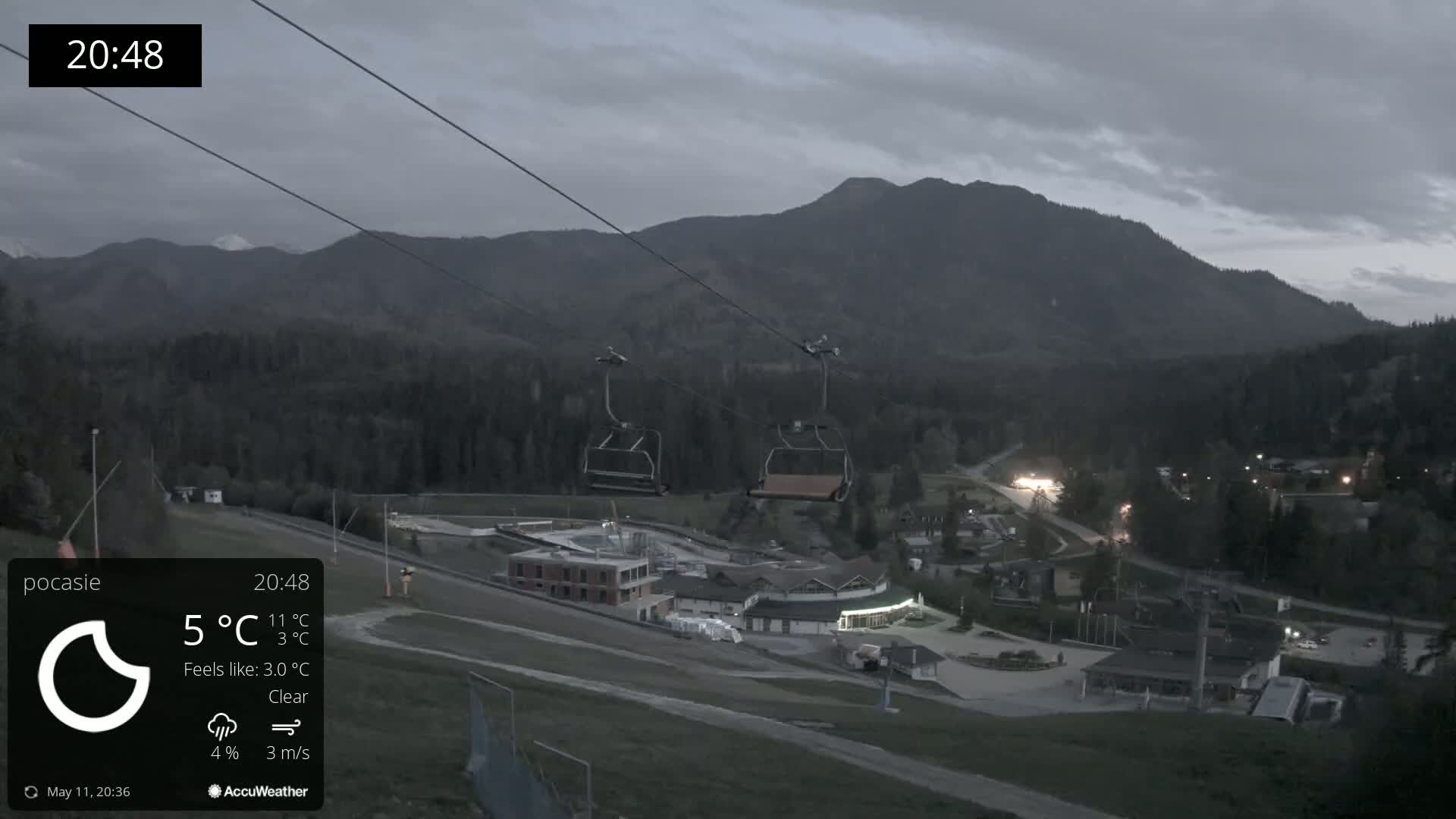 Under a clear, twilight sky, two empty chairlifts hang above a mountain resort nestled amongst dark, forested hills.