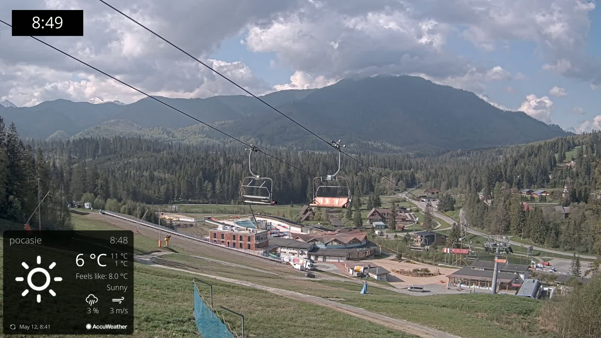 A sunny day at a ski resort shows an empty chairlift against a backdrop of mountains and a village below.