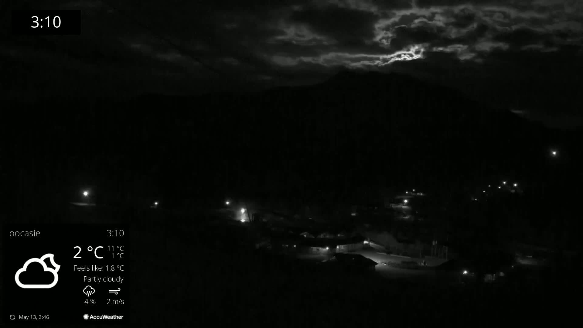 A nighttime, partly cloudy scene shows a small village nestled below a dark mountain under a sky with some bright clouds.