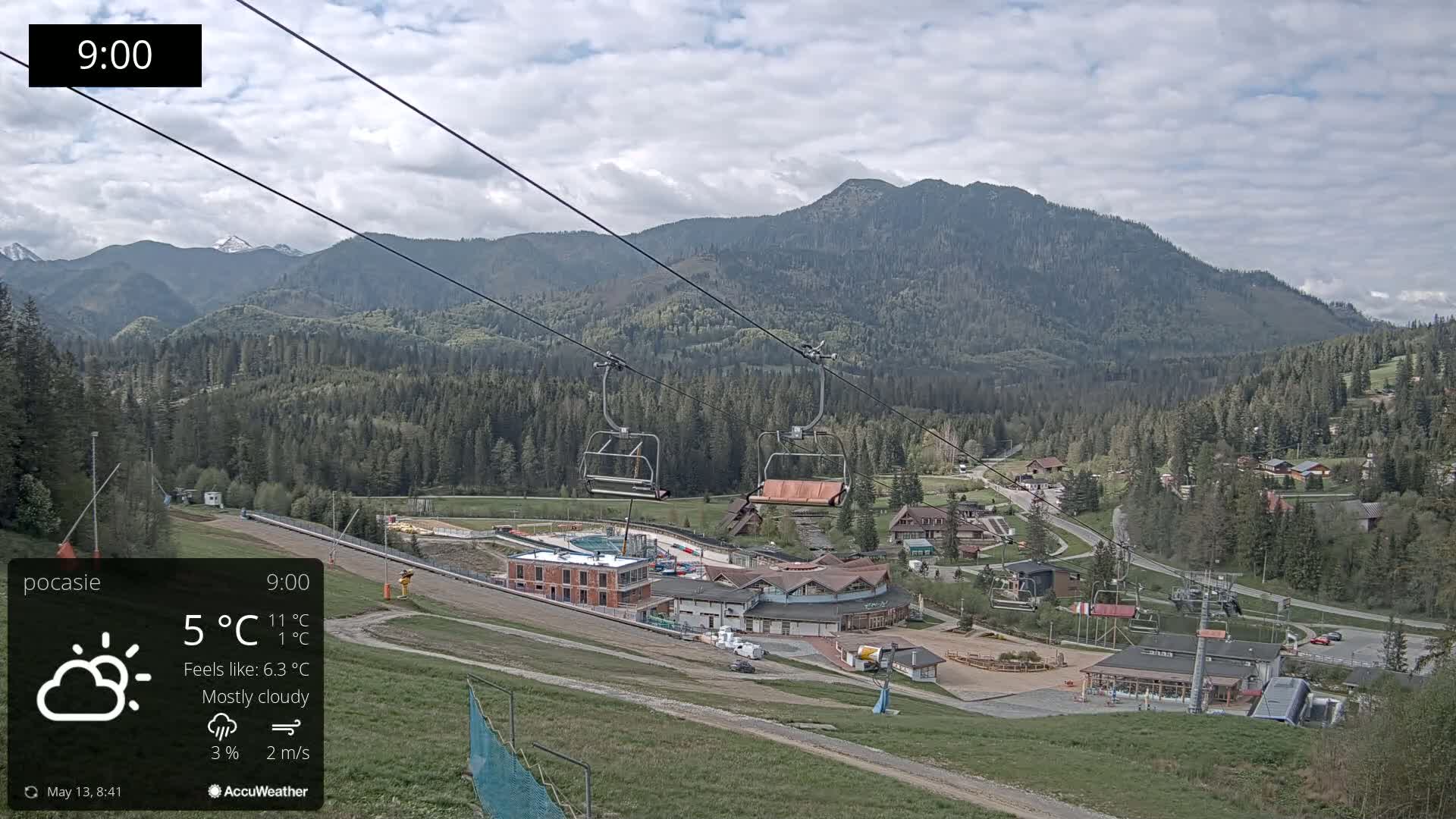 A mostly cloudy mountain valley scene with a chairlift and buildings, under a sky with some scattered clouds.