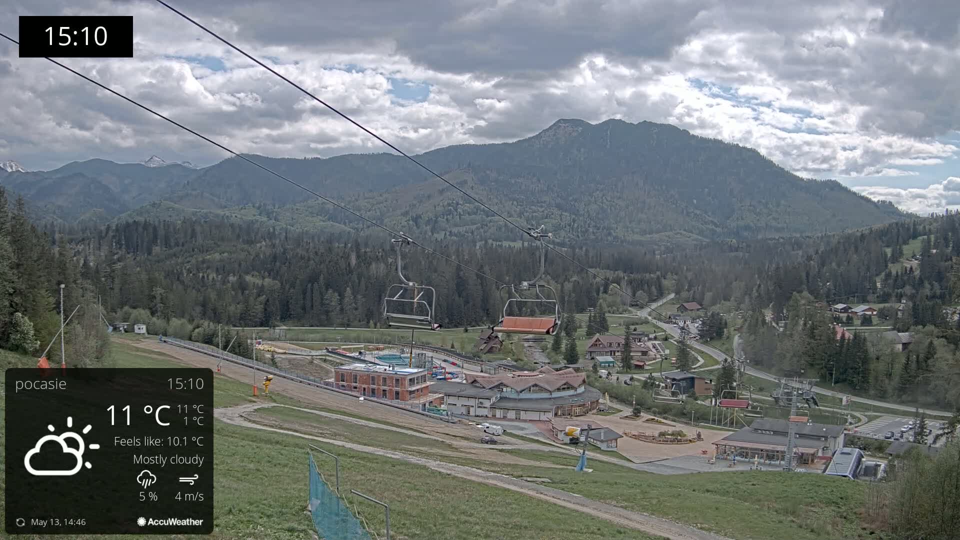 A mostly cloudy mountain scene with a chairlift, buildings, and a small town in the valley below.