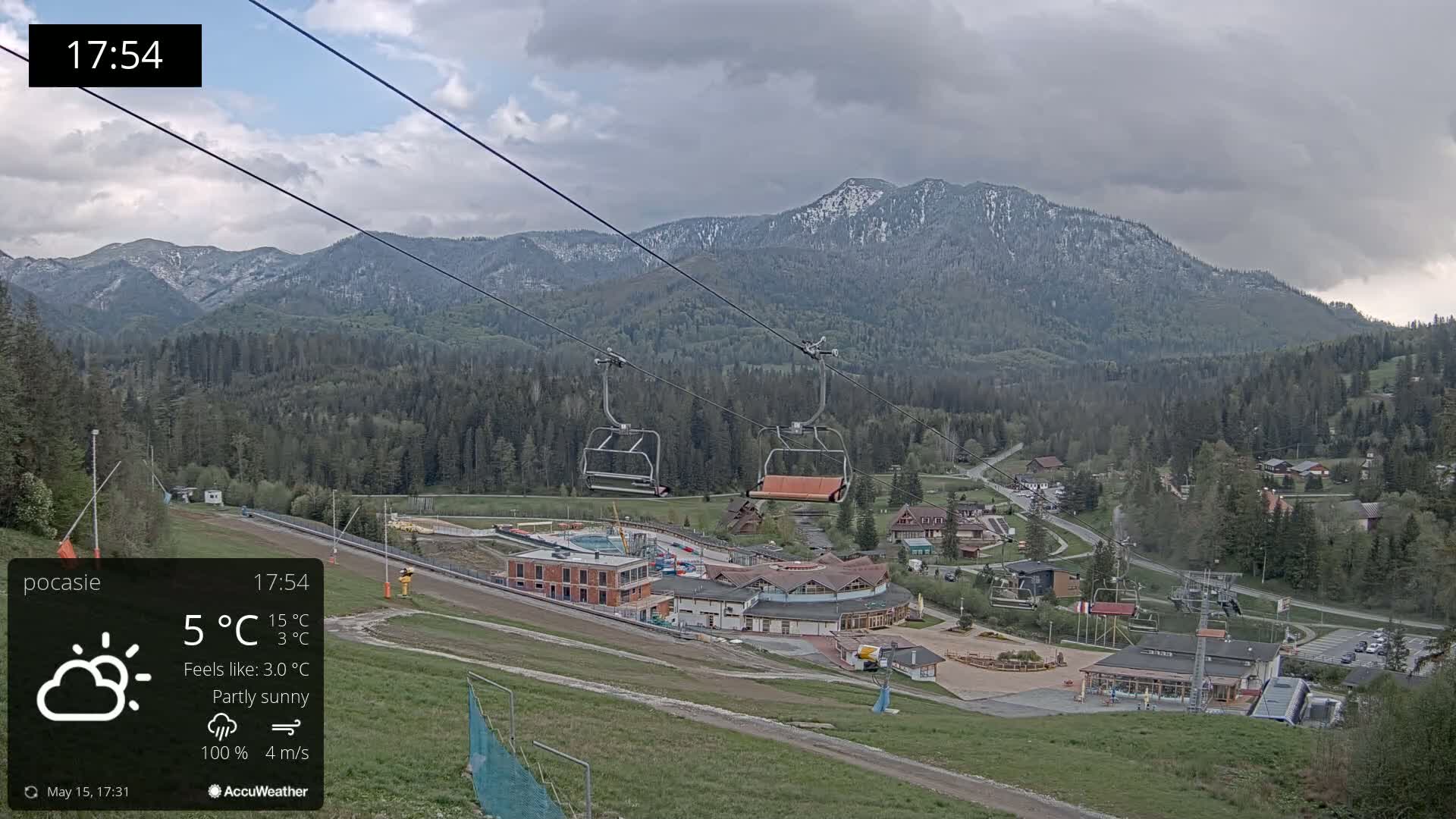 A partly sunny mountain vista shows a chairlift ascending towards a ski resort nestled among green trees and buildings, under a cloudy sky.