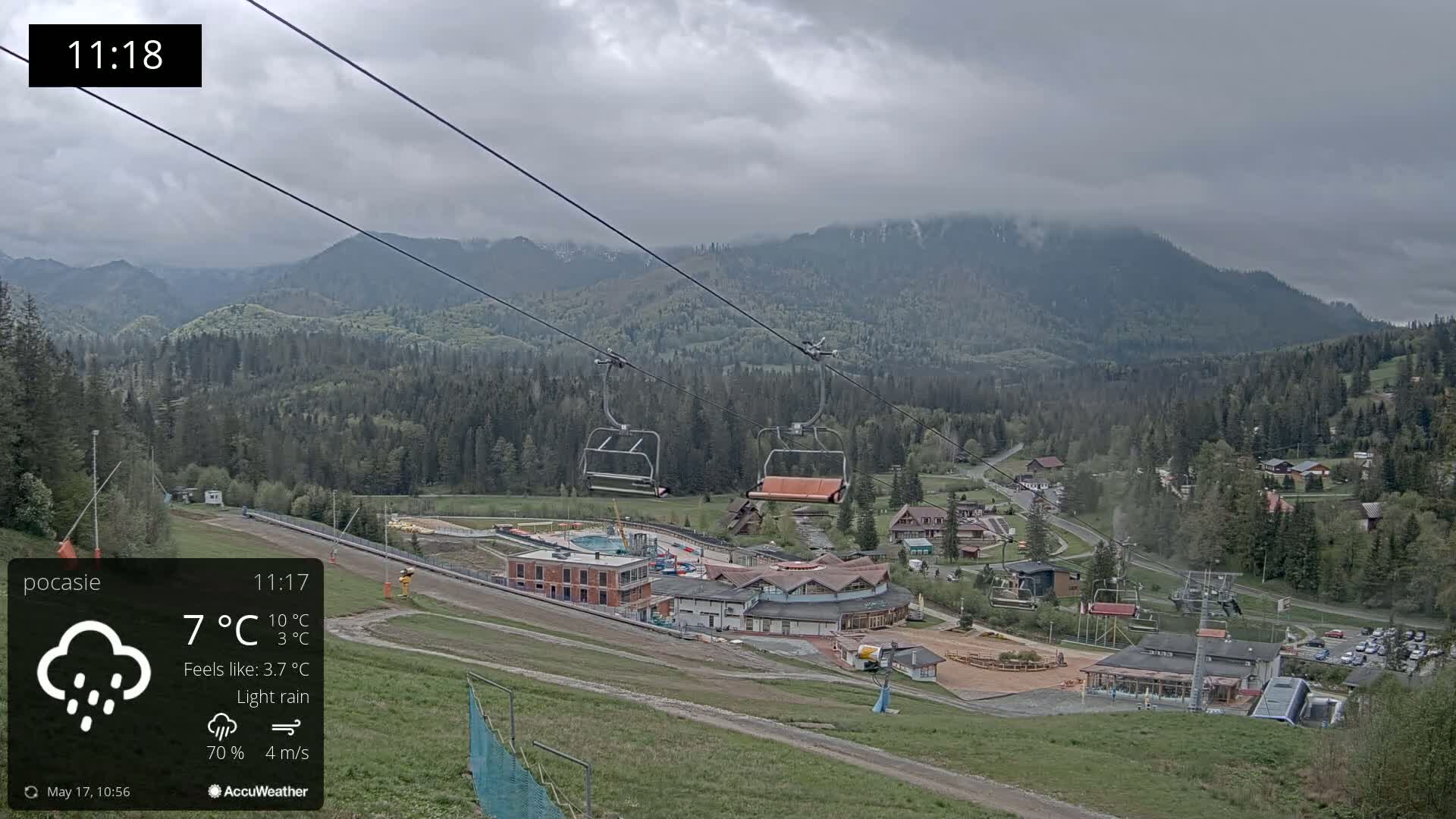 An overcast sky with light rain falls on a mountain resort, showcasing a chairlift and buildings nestled amongst green hills and forests.