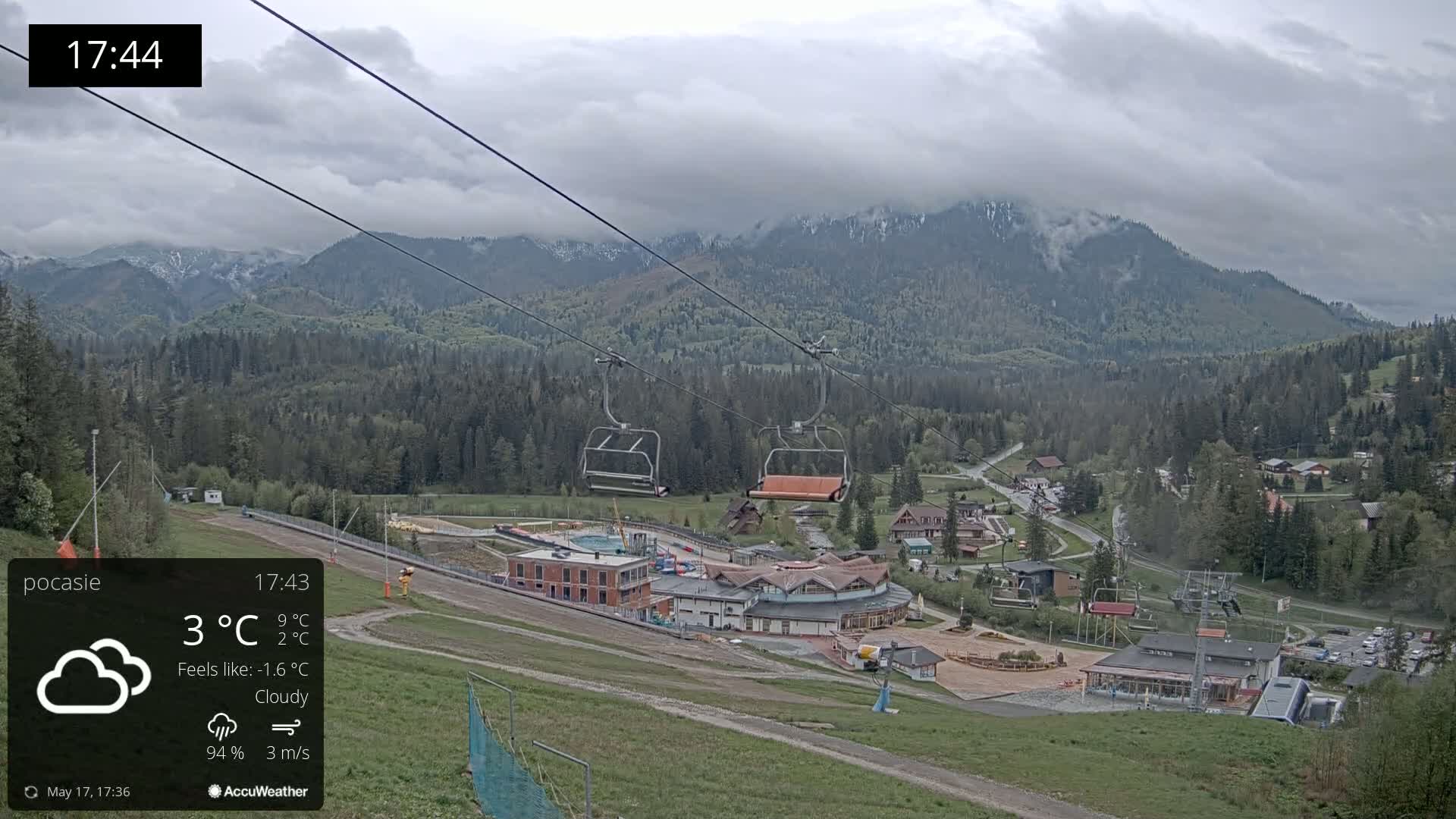 An overcast mountain valley scene features a chairlift ascending towards a resort, with low clouds partially obscuring the snow-capped peaks.