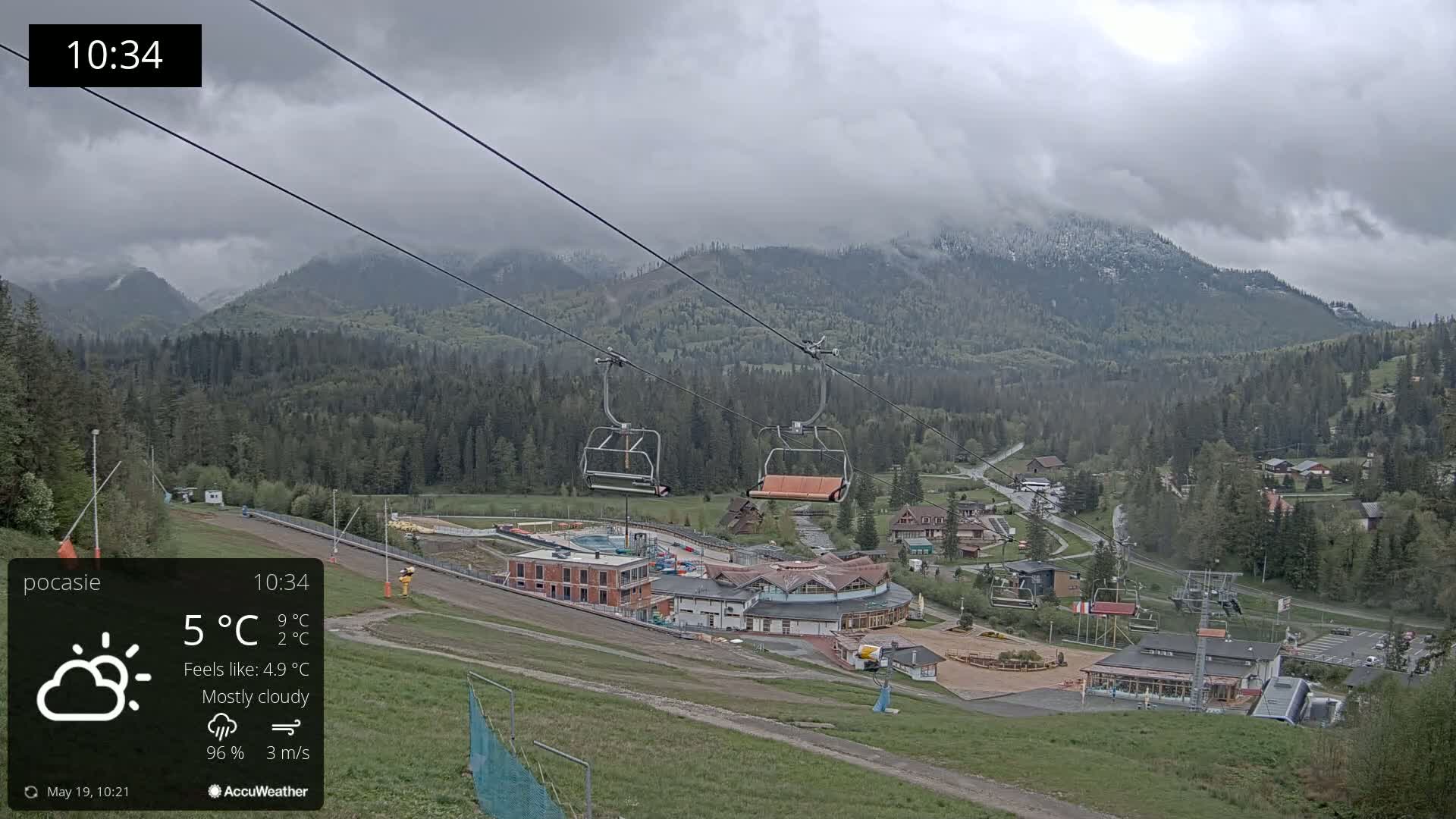 A mostly cloudy mountain scene with light snow on the peaks, showing a ski lift and resort buildings below under a grey sky.