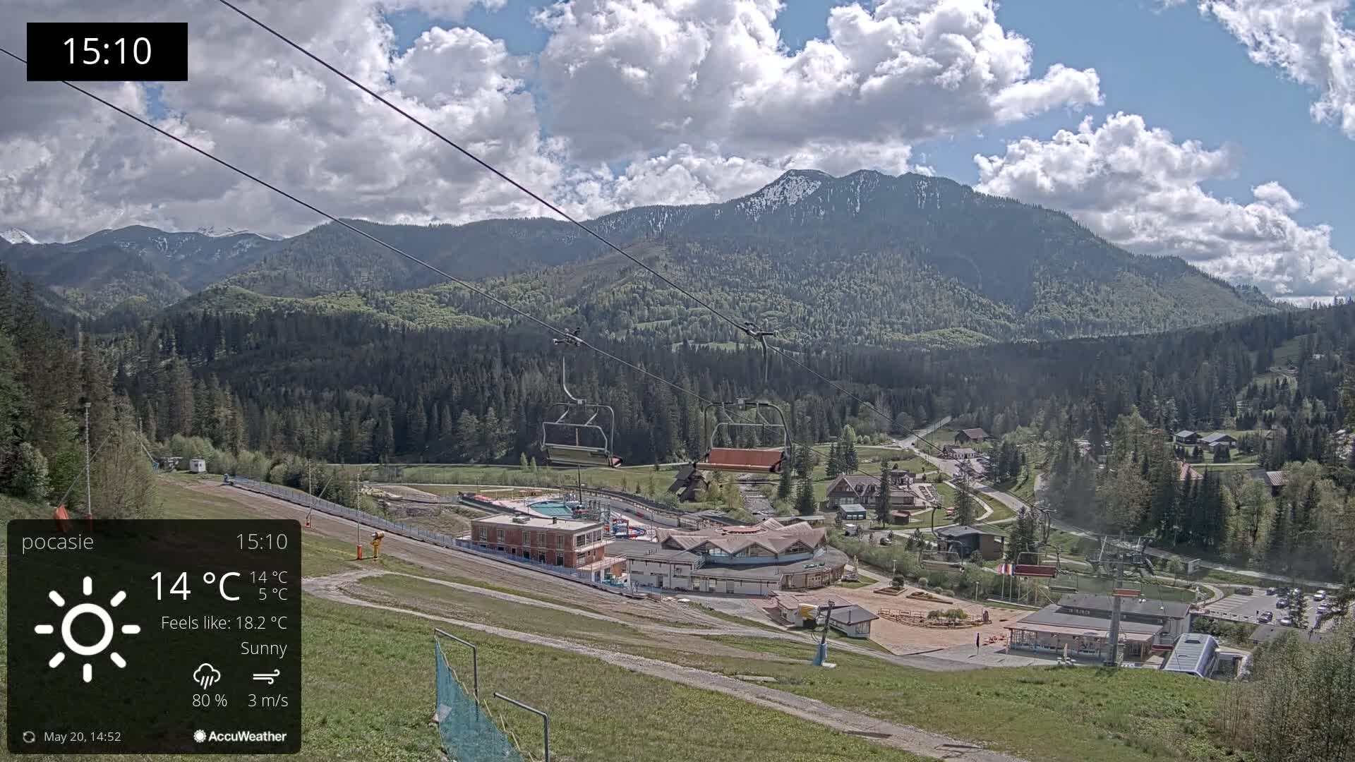 A sunny mountainside view shows a ski lift with empty chairs overlooking a resort village nestled among green trees and mountains under a partly cloudy sky.