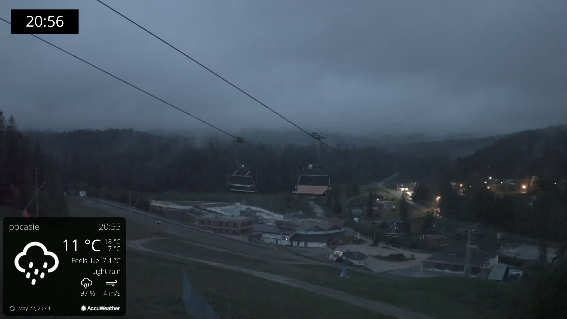 Two empty chairlifts ascend a hill overlooking a village under a dark, misty sky with light rain.