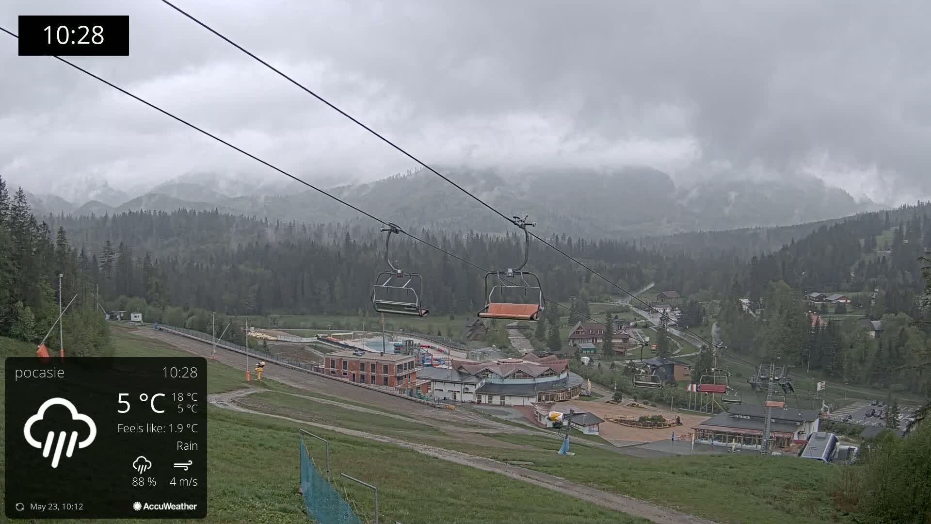 A rain-covered mountainside village with a chairlift and buildings is seen under a gray, cloudy sky.