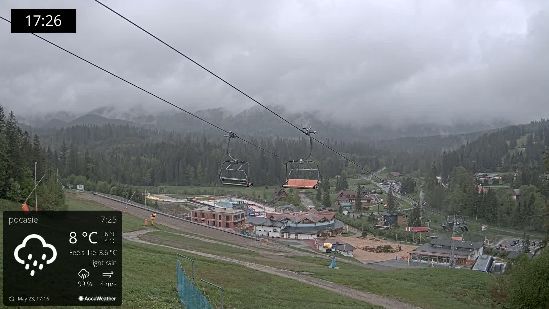 An overcast, lightly rainy mountain scene shows an empty chairlift ascending toward a ski resort nestled in a valley of green trees.
