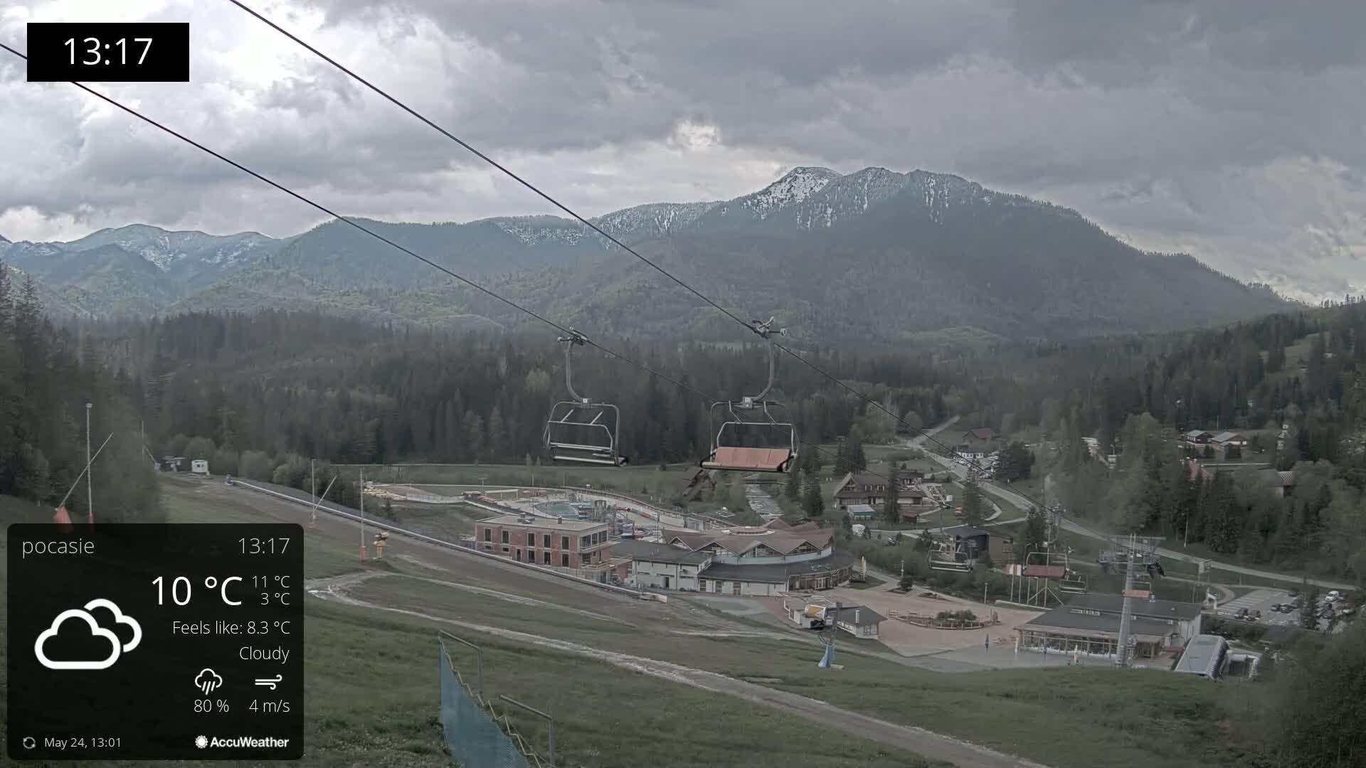 A chairlift ascends a mountainside toward a cloudy sky, overlooking a village nestled among green hills and partially snow-capped mountains.