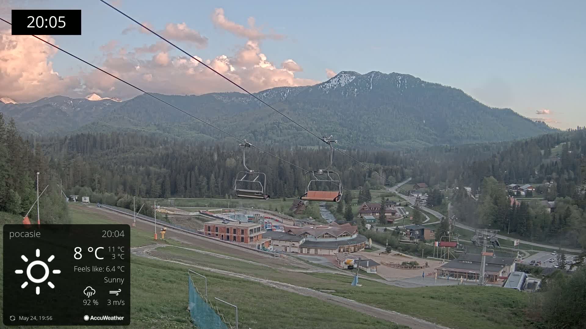 A sunny mountain resort with a chairlift, under a partly cloudy sky, overlooks a valley dotted with buildings and trees.