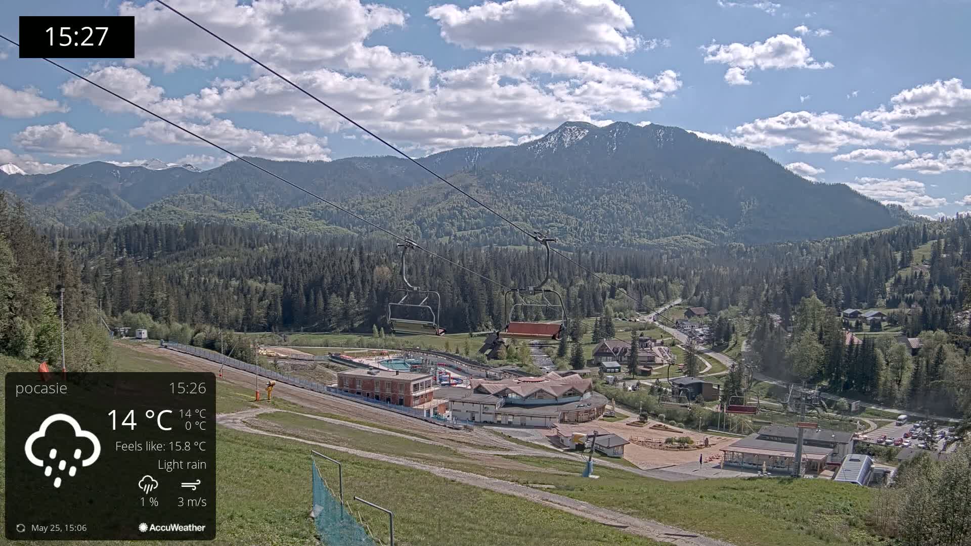 A chairlift ascends a mountainside overlooking a village nestled in a valley under a partly cloudy sky with light rain.